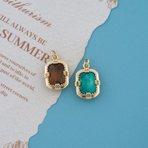 May include: Two rectangular pendants with gold-toned metal frames featuring decorative beaded edges and small loops at the top. One pendant contains a brown, textured stone, while the other contains a smooth, bright turquoise-colored stone.