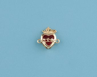 May include: A gold-colored heart-shaped charm with a red center, a crown, and a thorn design. The charm has two loops on either side. The background is a solid light blue.
