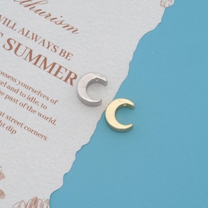 May include: Two small crescent moon charms, one in a polished silver tone and one in a gold tone, each featuring a small hole at the top for attachment. The charms are positioned on a textured white paper surface with printed text.