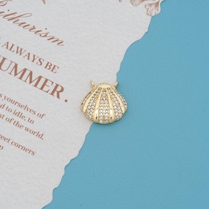Pode incluir: Um pingente de concha em metal dourado com uma série de sulcos verticais incrustados com pequenas pedras de zircônia cúbica transparentes e brilhantes. O pingente possui duas pequenas argolas no topo e está sobre um papel texturizado.