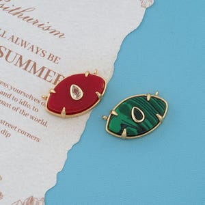 May include: Two oval-shaped pendants with gold-toned metal settings. One pendant features a solid red stone with a central teardrop-shaped clear crystal, while the other features a green malachite-patterned stone with a central dark teardrop.