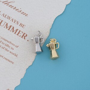 May include: Two small metal charms shaped like moka pot coffee makers, one in a silver tone and one in a gold tone. Each charm features a handle, a spout, and a small jump ring at the top for attachment, presented as miniature coffee brewing vessels.