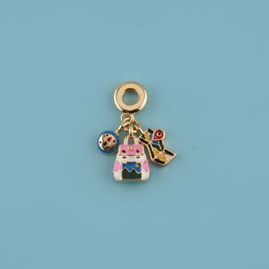May include: A gold-colored charm with a backpack, map, and compass design. The backpack is pink, white, blue, and green. The map is yellow, red, and black. The compass is blue and red. The charm is on a blue background.