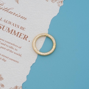 May include: A circular gold-toned hinged hoop ring featuring a row of small, clear, sparkling stones embedded along the outer edge. The jewelry piece has a smooth, polished finish and a secure clasp mechanism for opening and closing the hoop.