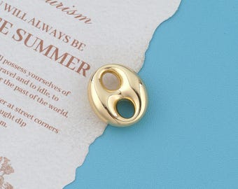 May include: A polished gold-tone metal bead with a smooth, rounded, oval-shaped design featuring two distinct circular openings in the center. The bead has a reflective surface and is positioned on a piece of paper with printed text.
