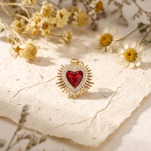 Puede incluir: Colgante de corazón dorado con una piedra central roja vibrante, rodeada de pequeños cristales transparentes. El colgante tiene un diseño de rayos de sol alrededor del corazón. Un pequeño lazo en la parte superior para una cadena. Joyería.