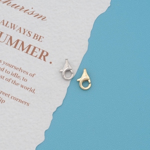 May include: Two lobster claw clasps, one in a silver tone and one in a gold tone, positioned next to a piece of white paper with the printed text "ALWAYS BE UMMER" and other partial words visible on the edge of the paper.