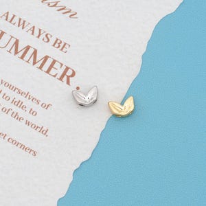 May include: Two small metal beads shaped like stylised leaves or wings, one in a silver tone and one in a gold tone. The beads are positioned on a piece of white paper with the printed text ALWAYS BE SUMMER and other partial words.