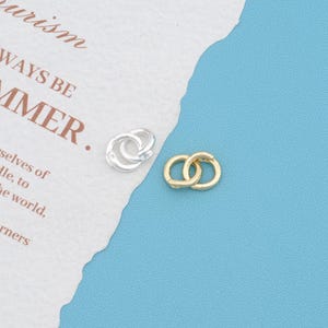 May include: Two small interlocking circular rings, one finished in a bright silver tone and the other in a polished gold tone. These metal components feature a smooth, rounded surface and are designed as individual jewelry findings or connectors.
