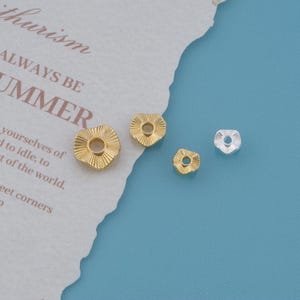 May include: Three gold-toned and one silver-toned heart-shaped beads with a textured, radial ribbed surface and a central circular hole. The beads vary in size, featuring a metallic finish and a distinct, pressed pattern on the front face.