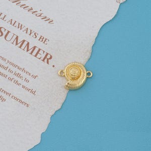 May include: A gold-toned metal charm shaped like a nautilus shell with a textured spiral center and two small loops on opposite sides for attachment. The charm features a polished finish and detailed ridges along the shell surface.