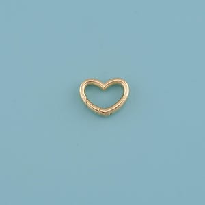 May include: A gold-colored heart-shaped clasp, likely for jewelry or crafts, is centered against a light blue background. The clasp is made of metal and has a hinged design for opening and closing.