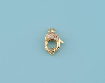 May include: A gold-colored lobster clasp with a decorative, rounded top encrusted with small, clear crystals. The clasp has a circular ring at the bottom and a small lever for opening and closing. The background is a solid light blue.
