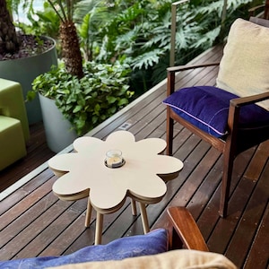 May include: A small, flower-shaped side table with a light wood finish. The table has a circular indentation in the center holding a small candle in a glass holder. The table is supported by three wooden legs. The table is on a wooden deck.
