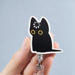 May include: A black cat badge reel with a white border and yellow eyes. The cat has a white design on its head. The badge reel has a clear plastic holder and a metal clip.