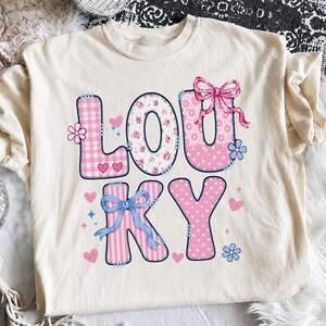 May include: Cream-colored t-shirt with the word "LOUKY" in a patchwork design. The letters are in pink, floral, and striped patterns, with hearts, bows, and flowers. The shirt has a crew neck and long sleeves.