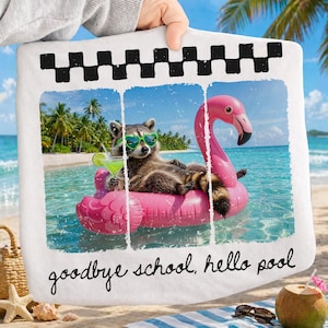 May include: A white beach towel with a photo-like print. The image shows a raccoon wearing sunglasses and floating on a pink flamingo pool float in the ocean. Text at the bottom reads "goodbye school, hello pool."