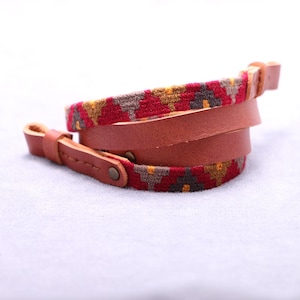 May include: A brown leather wrap bracelet with a woven red, yellow, and grey patterned band. The bracelet has a buckle closure and is coiled. The leather is a warm, medium brown colour.