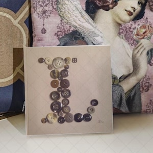 Peut inclure: Une carte carrée avec la lettre "L" composée de boutons de différentes tailles et couleurs. Les boutons sont disposés sur un fond beige clair avec un motif de losanges subtil. La carte est posée sur une surface blanche.