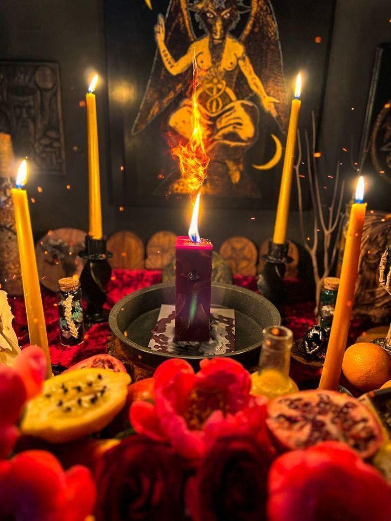 May include: A mystical scene featuring a central burning red candle with a tall flame, surrounded by lit yellow candles in black holders. The scene includes a red velvet surface, fruit, and a Baphomet image in the background.