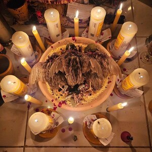 May include: An overhead shot of a ritualistic arrangement featuring a central dish with a bird, surrounded by lit candles and small wooden bowls. The scene is illuminated by the warm glow of the candles, with scattered red droplets.