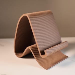 May include: A brown, ridged, curved tablet stand. The stand has a wave-like design with a back support and a lower lip to hold a tablet or phone. The stand is made of a solid material and has a matte finish.