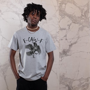 May include: A light gray t-shirt with the text "F-CAW-F" above a black illustration of a raven with outstretched wings. The raven is standing on one leg. The person is wearing a gold chain.