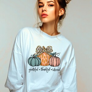 Peut inclure: Sweat-shirt blanc avec un motif de trois citrouilles turquoise, orange et rose, avec un nœud léopard. Le texte "grateful, thankful, blessed" est en dessous. Un design d'automne.