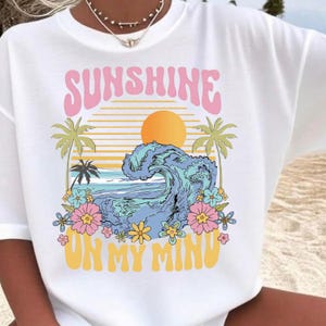 Peut inclure: T-shirt blanc avec un graphisme coloré. Le motif présente le mot "SUNSHINE" en rose, un soleil jaune, une vague océanique bleue, des palmiers et l'expression "ON MY MIND" en jaune.