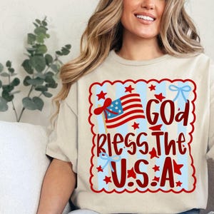 Könnte beinhalten: Beiges T-Shirt mit patriotischem Design. Das Design zeigt eine amerikanische Flagge, rote Sterne und den Text "God Bless The U.S.A." in Rot und Blau. Das Design ist mit einem roten, wellenförmigen Rand umrandet.