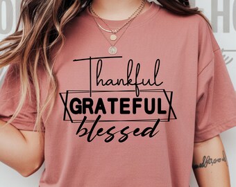 May include: A dusty rose-colored t-shirt with the words "Thankful, Grateful, Blessed" in black script. The word "Grateful" is inside a geometric frame. The shirt is a short-sleeved, crew neck style.