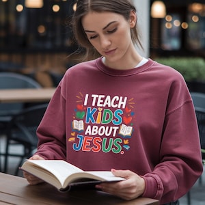 Peut inclure: Sweat-shirt bordeaux avec le texte "I TEACH KIDS ABOUT JESUS" en lettres et graphiques colorés. Le sweat-shirt a un col rond et des manches longues. Une personne tient un livre ouvert.