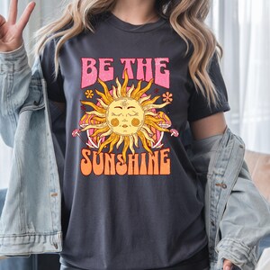 Peut inclure: T-shirt gris foncé avec un motif rétro. Il présente un soleil souriant aux yeux fermés, entouré de rayons et le texte "BE THE SUNSHINE" en rose et orange. Le t-shirt a une esthétique vintage et bohème.