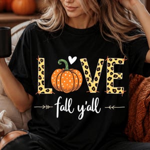 Peut inclure: Un t-shirt noir avec le mot LOVE en jaune à motif léopard, où la lettre O est remplacée par une citrouille orange à pois surmontée d'un cœur blanc, avec le texte fall y'all inscrit en dessous.