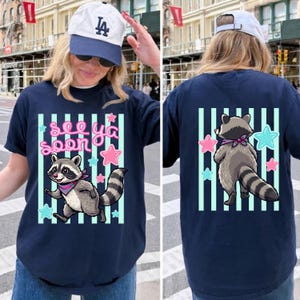 May include: Navy blue t-shirt with a raccoon graphic and the text "See Ya Soon". The front of the shirt features a raccoon with a pink bandana, surrounded by pink and blue stars. The back of the shirt also has a raccoon graphic.