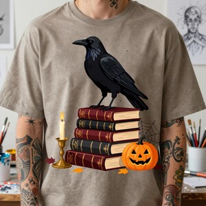 Peut inclure: Un t-shirt beige à manches courtes avec une impression graphique représentant un corbeau noir perché sur une pile de cinq livres. À côté des livres se trouvent une bougie allumée dans un bougeoir doré et une citrouille d'Halloween orange.