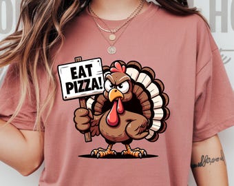 May include: A dusty rose-colored t-shirt featuring a cartoon turkey holding a sign that reads "EAT PIZZA!". The turkey has a stern expression, a red wattle, and a brown body with a fan of feathers. The shirt is a casual style.