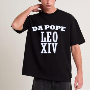Peut inclure: T-shirt noir avec l'inscription blanche "DA POPE LEO XIV". Le t-shirt a un col rond et des manches courtes. Le modèle porte un jean gris foncé.