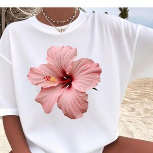 Peut inclure: T-shirt blanc orné d'un grand motif de fleur d'hibiscus rose. La fleur présente un centre rouge foncé et des étamines jaunes. Le t-shirt est de style ample et surdimensionné, idéal pour un look décontracté.