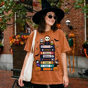 Peut inclure: Un t-shirt à manches courtes de couleur rouille avec une impression graphique représentant une pile de cassettes VHS étiquetées Hocus Pocus, Beetlejuice, Halloween, Friday the 13th, Casper et Coraline, entourées de personnages, chauves-souris et citrouilles.