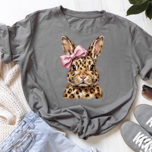 Peut inclure: T-shirt gris avec une illustration aquarelle d'un lapin. Le lapin a une fourrure à imprimé léopard et un nœud rose. La fourrure est un mélange de taches marron et beige. Le t-shirt est décontracté et confortable.