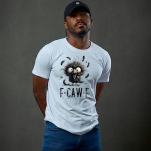 May include: A light blue t-shirt featuring a cartoon bird with large eyes and feathers. The text "F-CAW-E" is printed below the bird. The person in the image is wearing a black baseball cap and blue jeans.