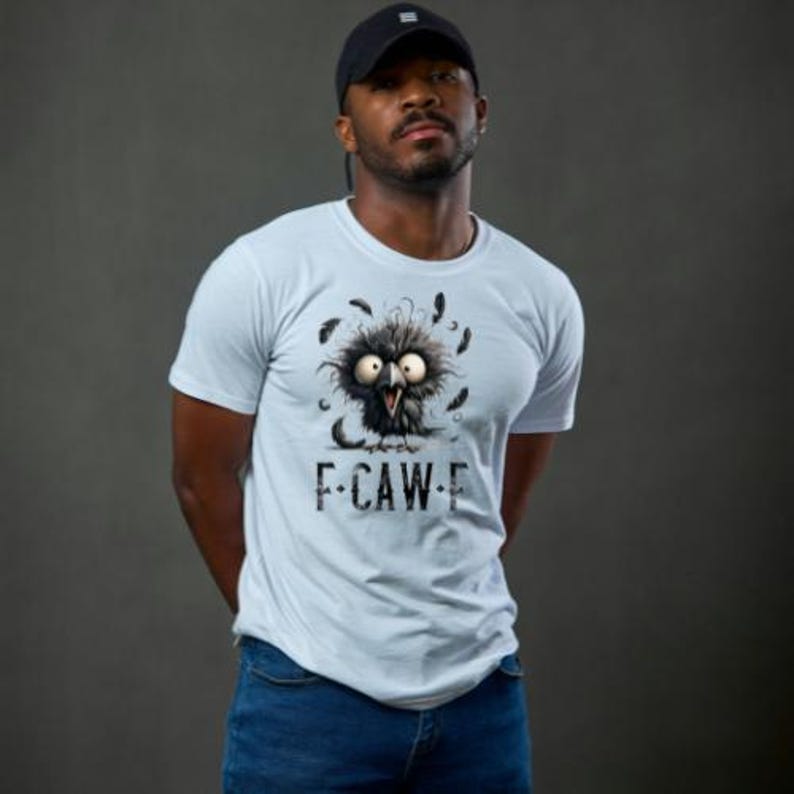 May include: A light blue t-shirt featuring a cartoon bird with large eyes and feathers. The text "F-CAW-E" is printed below the bird. The person in the image is wearing a black baseball cap and blue jeans.