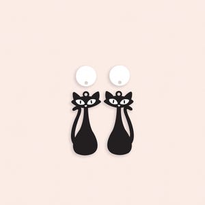 May include: Black cat-shaped earrings with white accents and a matching pair of white circular earring studs. The earrings have a minimalist design, featuring a sleek, elongated body and a small loop for hanging. The background is a soft, pale pink.