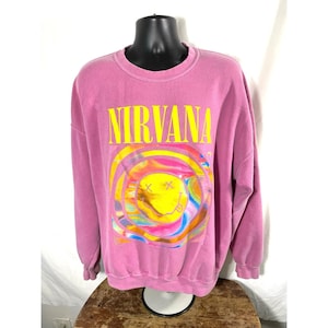 May include: A pink sweatshirt with the word "NIRVANA" in yellow block letters. Below the text is a colorful graphic of a smiley face with swirly rainbow colors. The sweatshirt is displayed on a mannequin.