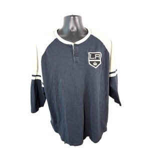 May include: A dark navy blue and white henley shirt featuring three-quarter length sleeves with two horizontal white stripes on each arm. The chest displays a shield-shaped patch with the letters LA and a crown graphic above the text.