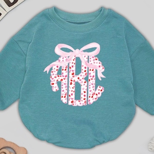 May include: Teal baby romper with a pink bow and monogram design. The monogram features the letters "ABH" filled with red and pink hearts. The romper has long sleeves and a round neckline. Wooden blocks spell out "BABY" in the upper left corner.