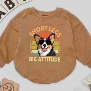 May include: A brown baby onesie with long sleeves. The onesie features a graphic of a corgi wearing sunglasses with the text "SHORT LEGS BIG ATTITUDE". The word "BABY" is spelled out in wooden letters above the onesie.