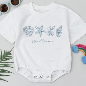 May include: White baby bodysuit with a blue seashell design and the name "William" printed below. The bodysuit has a round neck and snap closures. Sunglasses and a rainbow decoration are also in the image.