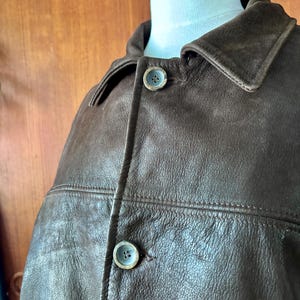 Peut inclure: Une veste en cuir marron foncé dotée d'un col classique, d'une patte de boutonnage avant avec deux boutons circulaires visibles et de coutures horizontales sur la poitrine. Le vêtement est présenté sur un buste de mannequin blanc.